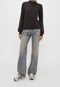Person trägt einen schwarzen langen Rollkragenpullover, hellblaue weite Jeans und weiße Sneakers mit schwarzen Details und steht vor einem schlichten Hintergrund.