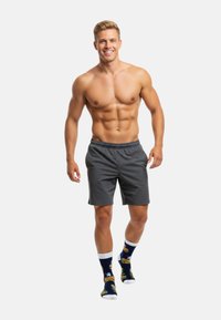 Grå atletiske shorts, mønstrede sokker med burgere og en tegneseriefigur, båret af en smilende mand med muskuløs kropsbygning.