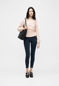 Γυναίκα που φοράει ροζ πουκάμισο με ρυτίδες, σκούρο στενό παντελόνι, μαύρα μοκασίνια και κρατά μια μεγάλη μαύρη τσάντα ώμου, στέκεται μπροστά σε ένα απλό φόντο.