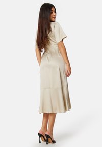 Robe en satin beige à manches courtes, taille cintrée et ourlet évasé. Associée à des mules à talons noirs, montrant une texture lisse et un drapé élégant.