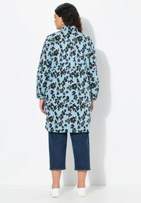 Camicia a maniche lunghe di colore azzurro chiaro con un motivo astratto nero e verde. Vestibilità ampia, colletto alto e orlo arrotondato. Abbinata a pantaloni in denim a vita alta.
