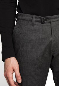Pantaloni grigi textured con frontale liscio, chiusura con bottone e tasche laterali. Indossati con una camicia nera a maniche lunghe, che mette in evidenza una vestibilità slim.