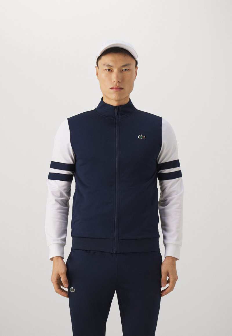 Veste zippée marine avec des manches blanches, présentant des rayures horizontales bleues nuit et un logo vert sur la poitrine gauche. Tissu doux, coupe athlétique.