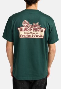 Camiseta de algodón verde con un gráfico en la parte posterior que incluye una rosa y el texto: "Balance de Opuestos, Costa Mesa, CA, Servicio y Pantalones."