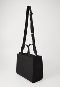 Sac fourre-tout en toile noir avec deux poignées supérieures et une bandoulière réglable. Présente une forme structurée et une poche avant. Texture lisse.