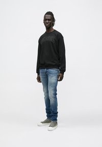 Νέος άνδρας που φοράει μαύρο φούτερ Diesel, ξεθωριασμένο μπλε τζιν και γκρι ψηλά αθλητικά παπούτσια, στέκεται μπροστά σε απλό λευκό φόντο.