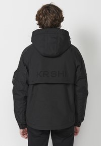 Svart huva jacka med dragkedja, elastiska ärmslut och en distinkt perforerad logotyp på baksidan. Har en avslappnad passform.