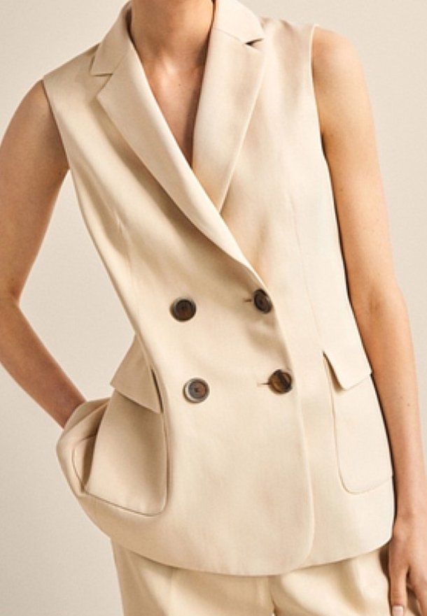 Blazer beige sans manches à double boutonnage avec quatre boutons foncés et poches à rabat, porté avec un pantalon assorti.