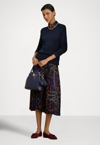 Nő sötétkék pulóverben és mintás, pliszírozott szoknyában, tengerészkék kézitáskát tartva, bordó loafereket és aranylánc nyakláncot viselve, fehér háttér előtt áll.