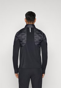 Μαύρο αθλητικό μπουφάν με γέμιση από πούπουλο στην επάνω του περιοχή, μακριά μανίκια και ψηλό γιακά. Ο πίσω σχεδιασμός περιλαμβάνει ανακλαστική λεπτομέρεια και υφή υφάσματος.