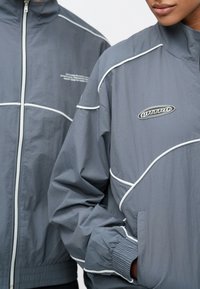 Grå hettejakker med glidelås og hvite pipedetaljer. Begge har et teksturert, glatt stoff og ribbestrikkede mansjetter, med en liten logo på brystet.