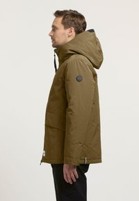Olijfgroene geïsoleerde jas met een capuchon, een ritssluiting aan de voorkant, een grote voorzak en een cirkelvormig logo op de bovenarm. Glad, matte textuur.