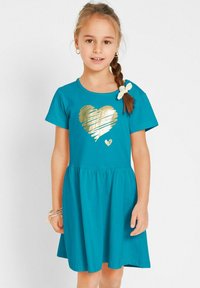 Robe en coton teal avec un design à manches courtes, ornée d'un graphique en forme de cœur doré et d'une jupe à volants. Un accent en forme de nœud blanc décore l'épaule.