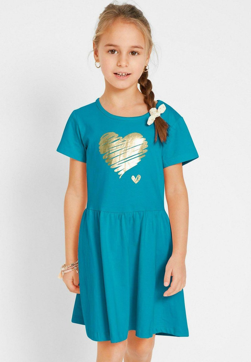 Robe en coton teal avec un design à manches courtes, ornée d'un graphique en forme de cœur doré et d'une jupe à volants. Un accent en forme de nœud blanc décore l'épaule.