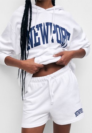 Person mit langen Zöpfen hebt einen weißen Hoodie mit dem Text "NEWPORT" und trägt passende weiße Shorts mit Kordelzug und "NEWPORT" am Bein.