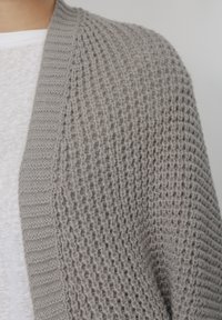 Κοντινή λήψη γκρι πλεκτής ζακέτας με υφή μοτίβου, που φοριέται πάνω από ένα λευκό πουκάμισο.