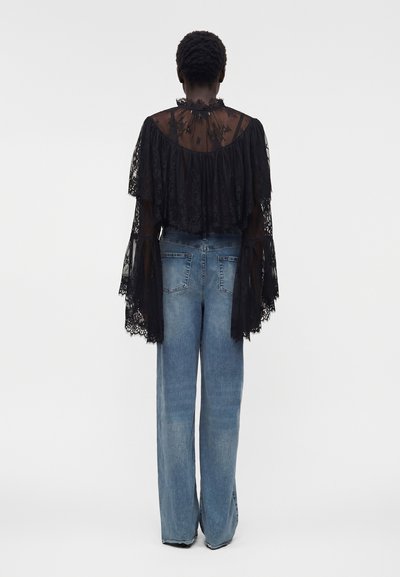 Άτομο με διάφανο μαύρο δαντελωτό μπλουζάκι με μακριά φαρδιά μανίκια και ψηλόμεσα μπλε jeans με ίσια μπατζάκια, στραμμένο πλάτη προς τον θεατή.