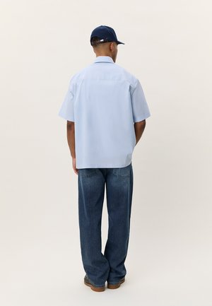 Άνδρας που φοράει ανοιχτό μπλε κοντομάνικο πουκάμισο, σκούρο μπλε τζιν, καφέ παπούτσια και σκουρό μπλε καπέλο, στέκεται με την πλάτη προς τα εμπρός σε απλό φόντο.