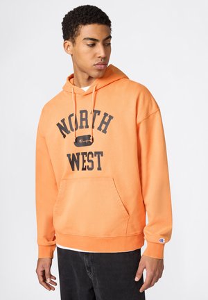 Młody mężczyzna w pomarańczowej bluzie z kapturem "North West" z przednią kieszenią i czarnych spodniach, patrzący w lewo na jednolitym tle.