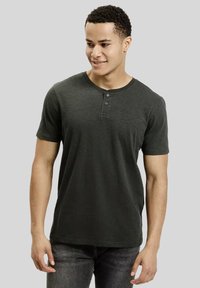 Młody mężczyzna z krótkimi, kręconymi włosami, ubrany w ciemnoszary krótki t-shirt henley i ciemne dżinsy, patrzy nieco w bok, uśmiechając się.