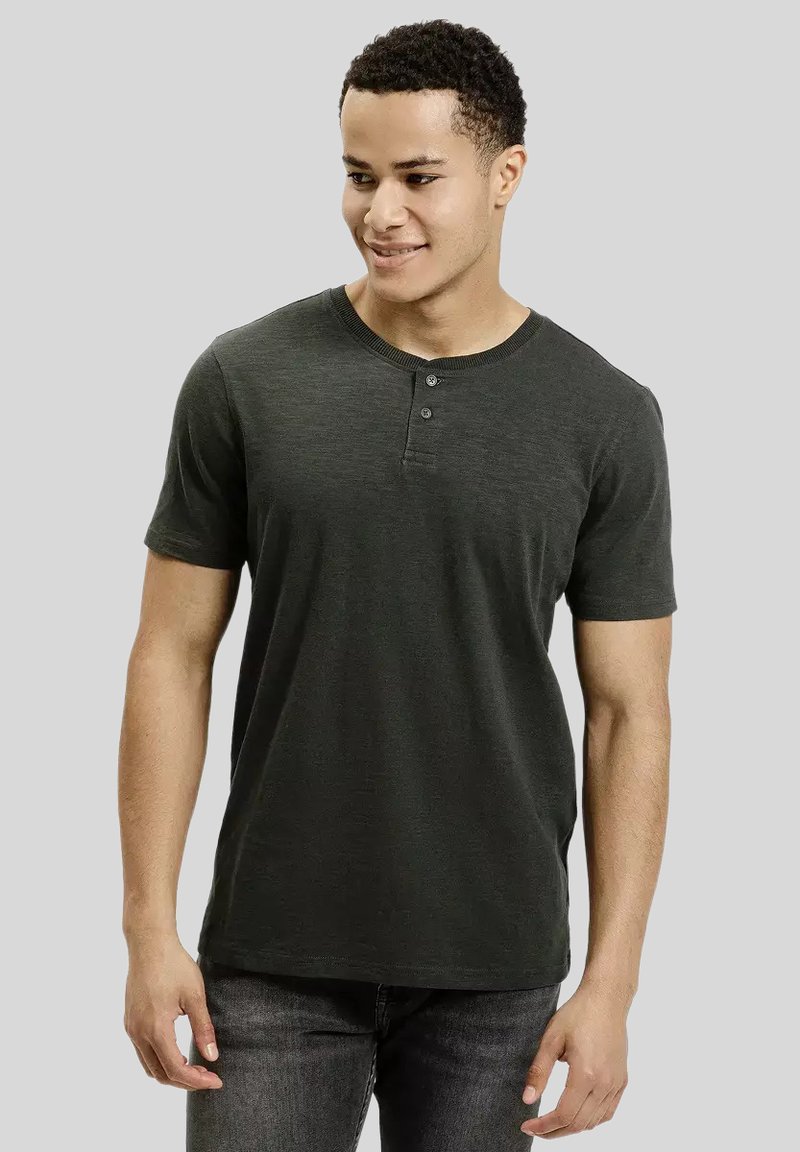 Młody mężczyzna z krótkimi, kręconymi włosami, ubrany w ciemnoszary krótki t-shirt henley i ciemne dżinsy, patrzy nieco w bok, uśmiechając się.