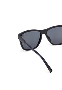 Occhiali da sole neri con lenti grigie, caratterizzati da un design angolare elegante. Aste nere texturizzate con linee di accento blu e dettagli del logo.