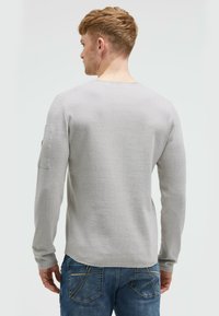 Hellgraues Langarmhemd mit strukturiertem Oberflächen, rundem Halsausschnitt und einer aufgenähten Tasche am linken Ärmel. Getragen mit blauen Jeans.