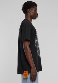 Black cotton t-shirt with a large graphic of a skull design in white and blue. Features a rectangular orange tag at the hem.