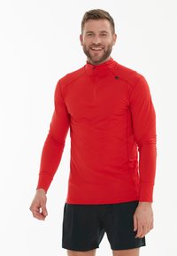 Rød langærmet skjorte med høj krave og halv-zip design, kombineret med sorte shorts. Stofdet ser glat og tætsiddende ud.