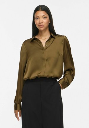 Femme aux longs cheveux foncés portant un chemisier en satin vert olive rentré dans un pantalon noir taille haute, debout avec une main dans la poche.