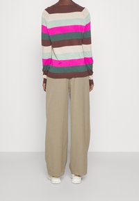 Henkilö, joka on pukeutunut leveisiin beigenvärisiin housuihin, valkoisiin lenkkareihin ja pitkähihaiseen neuleeseen, jossa on vaaka raidat pinkkiä, ruskeaa, vihreää ja beigeä, selin katsojaan.