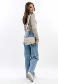 Beige crossbody taske med en tekstureret overflade, lynlåslomme foran og justerbar rem, holdt af en person iført afslappet påklædning.