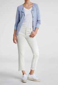 Un cardigan bleu clair sur un T-shirt blanc à col en V, associé à un jean blanc coupé avec un ourlet effiloché et des détails de boutons. Terminé avec des baskets blanches.
