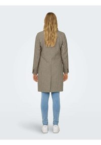 Cappotto marrone a scacchi con maniche lunghe, taglio dritto e spacco posteriore, abbinato a jeans skinny blu chiaro e sneakers bianche.
