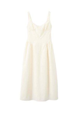 Frau mit lockigem Haar, die ein ärmelloses weißes Kleid und schwarze Absätze trägt, steht mit dem Gesicht zu einer einfarbigen roten Wand und hält eine schwarze Handtasche.