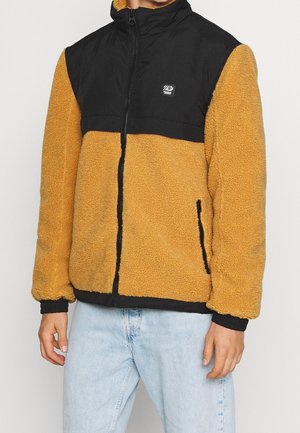 Man met een beige en zwarte fleecejas met ritssluiting aan de voorkant en lichtblauwe spijkerbroek tegen een eenvoudige achtergrond.