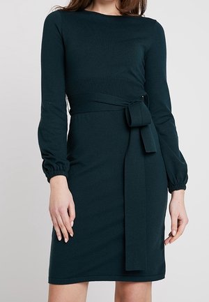 Vrouw draagt een donkergroene jurk met lange mouwen, een aangesloten taille en een geknoopte stoffen ceintuur, staand tegen een effen lichte achtergrond.