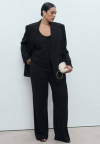 Frau mit dunklem Haar in einem glatten Dutt trägt einen schwarzen oversized Blazer, eine geknöpfte Weste, weite Hosen und hält eine kleine weiße geflochtene Clutch.