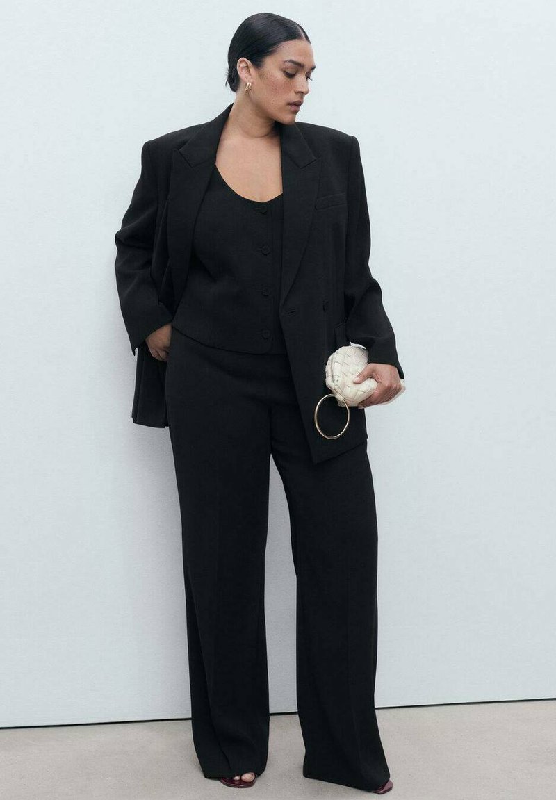 Frau mit dunklem Haar in einem glatten Dutt trägt einen schwarzen oversized Blazer, eine geknöpfte Weste, weite Hosen und hält eine kleine weiße geflochtene Clutch.