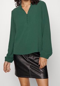 Grön långärmad topp med v-ringning och fem knappslitsar, kombinerad med en svart läder minikjol med kontrasterande sömnadsdetaljer.