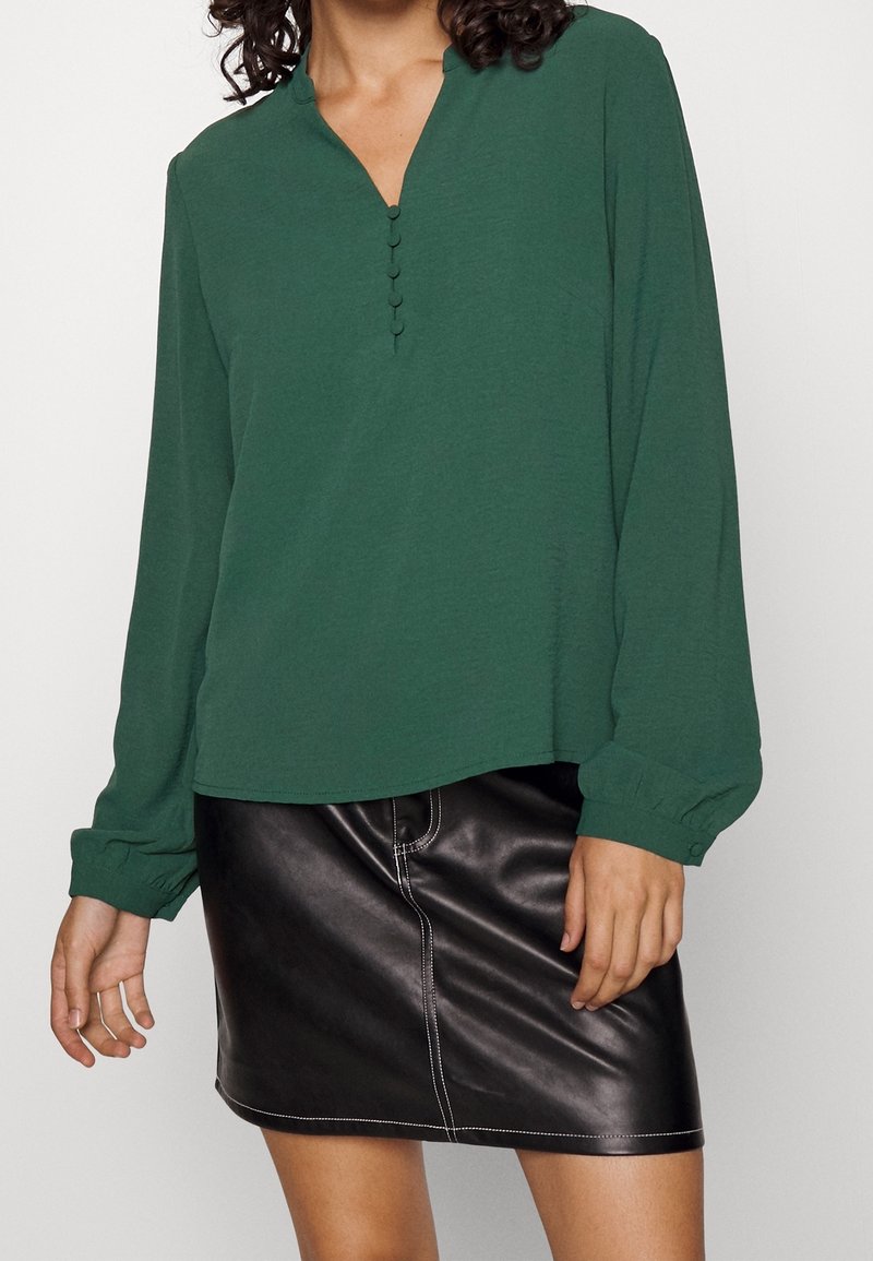 Grön långärmad topp med v-ringning och fem knappslitsar, kombinerad med en svart läder minikjol med kontrasterande sömnadsdetaljer.