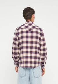 Camisa de cuadros en colores crema, azul y rojo; tela de algodón con un patrón a cuadros; puños con botones y detalle de canesú en la parte posterior.