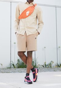 Hnědé košilové tričko přes béžové kraťasy, oranžová ledvinka Nike, černé a růžové tenisky a černé ponožky s bílými akcenty v neformálním prostředí.