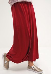 Pantaloni larghi rossi realizzati in un tessuto morbido, con una cintura elastica. Abbinati a scarpe slip-on dorate metalliche con una superficie testurizzata.