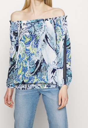 Vrouw draagt een off-shoulder blouse met een blauw, groen en wit abstract patroon, gecombineerd met lichtblauwe spijkerbroek.