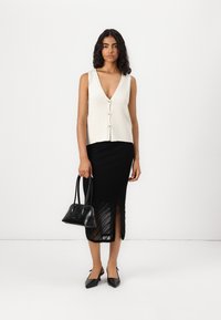 Kremowy, dzianinowy top na ramiączkach z dekoltem w kształcie litery V i zapięciem na guziki, zestawiony z czarną spódnicą do kolan z pionowymi paskami i rozcięciem.