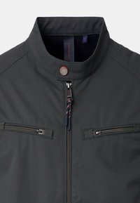 Chaqueta de color verde oscuro con un cuello alto, dos bolsillos en el pecho con cremallera y un cierre frontal con cremallera. Presenta costuras en contraste y un tejido texturizado.