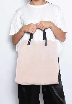 Sac fourre-tout en suède rose pâle avec des poignées en cuir noir et des coutures blanches. Forme rectangulaire simple, texture douce et sans matériel visible.