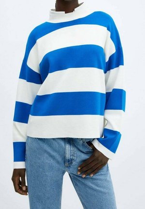 Person trägt einen weißen Pullover mit langen Ärmeln und breiten blauen horizontalen Streifen, kombiniert mit hellblauen Jeans, eine Hand in der Tasche.