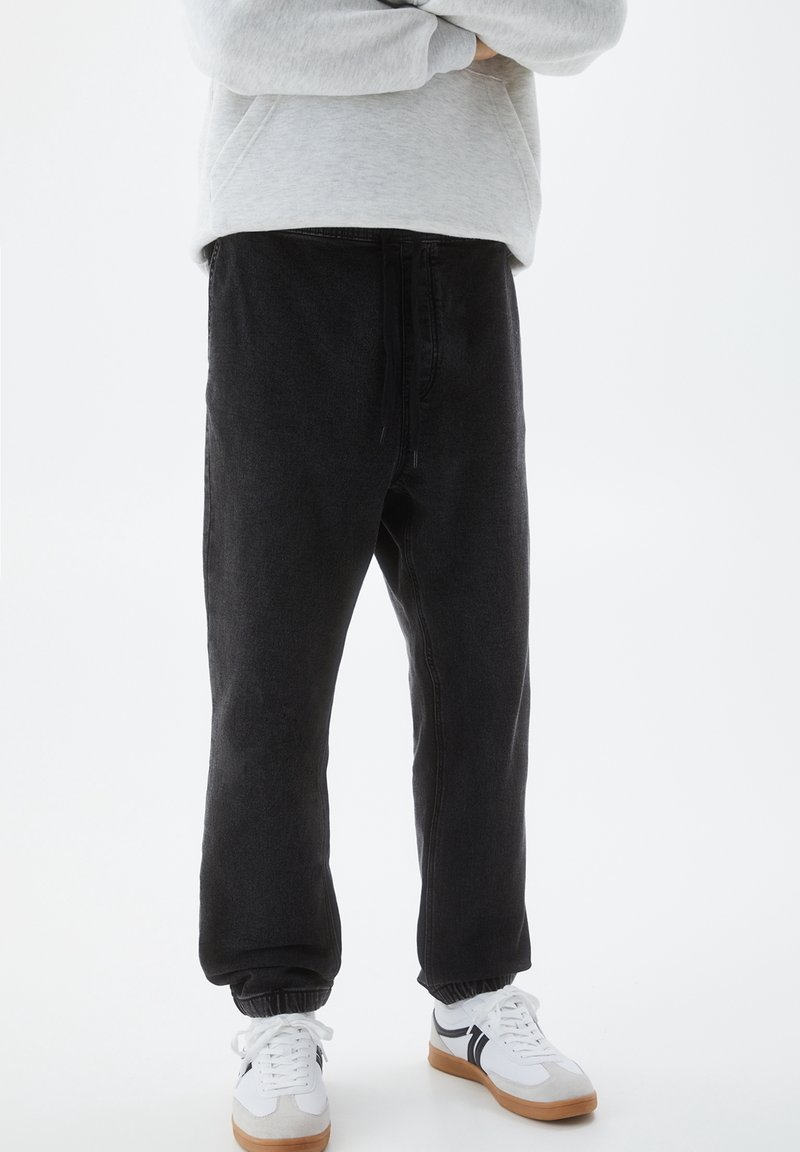 Pantaloni jogger neri realizzati in tessuto morbido, con cintura elastica e polsini. Indossati con sneakers bianche e una felpa grigia chiara.